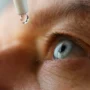 Primer plano de un ojo humano azul recibiendo una gota de colirio desde un gotero, con enfoque médico y luz natural que resalta el detalle y la precisión del tratamiento ocular.