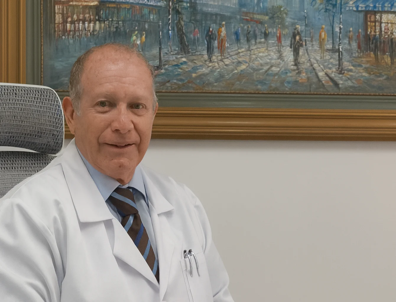 Retrato del Dr. Dagoberto Gallego Zapata con bata médica, sentado en su consultorio frente a un cuadro artístico.