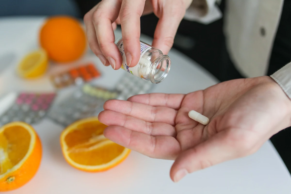 Persona tomando una cápsula de suplemento nutricional junto a naranjas y diferentes vitaminas